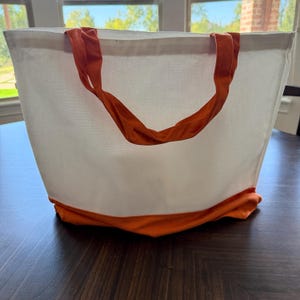May include: A white tote bag with orange accents. The bag features orange handles and a matching orange base. The bag is made of a sturdy fabric and is designed for carrying items. The bag is sitting on a dark wooden surface.