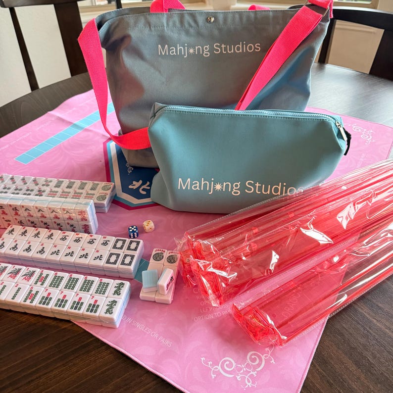 May include: A Mahjong set featuring a grey tote bag and a matching pouch, both with pink accents and the text "Mahjong Studios." The set includes tiles, dice, and red plastic tile racks, arranged on a pink mat.