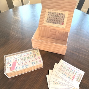 May include: A wooden box with an open lid, a clear acrylic case filled with tiles, and several cards. The box and case contain tiles for the game Mahjong. The box has a wood grain pattern and a printed label inside the lid.
