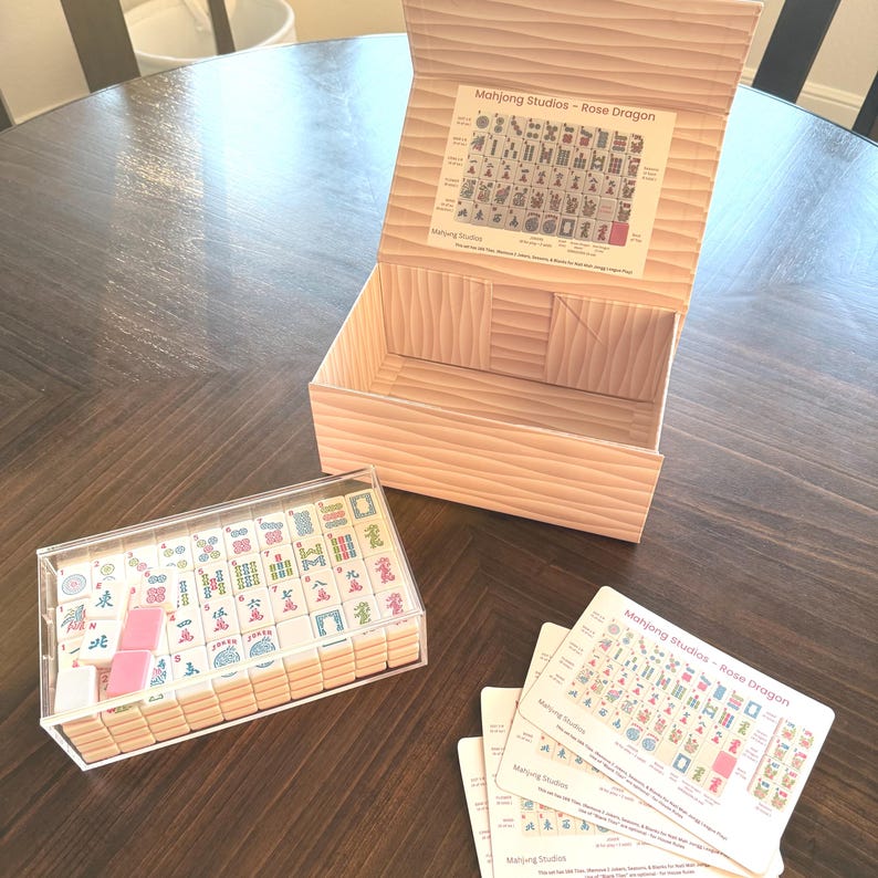 May include: A wooden box with an open lid, a clear acrylic case filled with tiles, and several cards. The box and case contain tiles for the game Mahjong. The box has a wood grain pattern and a printed label inside the lid.