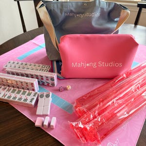 May include: A Mahjong set with a pink and gray theme. The set includes tiles, dice, and a pink mat. A gray tote bag and a pink zippered pouch, both with the text "Mahjong Studios", are also included. Red plastic sticks are visible.