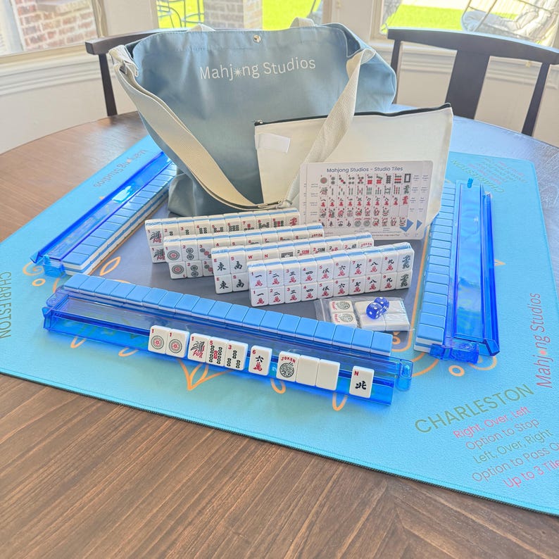 May include: A complete Mahjong set with blue accents, including tiles, racks, and a game mat. The set includes a light blue tote bag with "Mahjong Studios" printed on it. The game mat has the word "Charleston" printed on it.