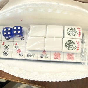 May include: A white zippered pouch containing a Mahjong set. The set includes white tiles with various symbols and the word "JOKER", and two blue dice. The tiles are packaged in clear plastic.