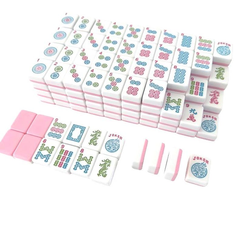 May include: A complete Mahjong set with white tiles featuring pink edges and colorful symbols. The set includes tiles with numbers, characters, and Jokers, arranged in stacks and rows. The game pieces are displayed on a white surface.
