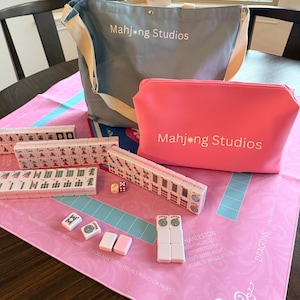 May include: A Mahjong set with a pink and blue game mat, tiles, dice, and a gray tote bag and pink pouch, both with "Mahjong Studios" printed on them. The tiles are white with red, green, and black markings. The game mat has text and a blue and pink design.
