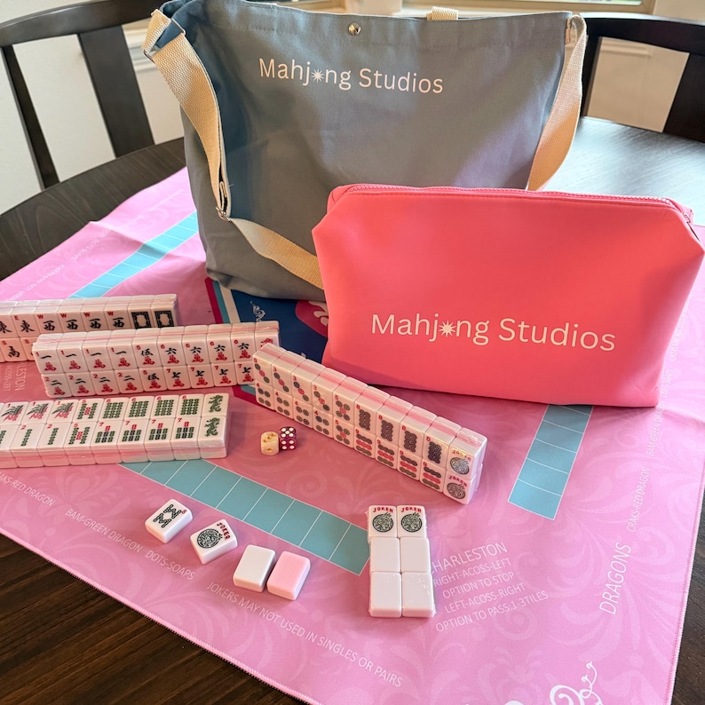 May include: A Mahjong set with a pink and blue game mat, tiles, dice, and a gray tote bag and pink pouch, both with "Mahjong Studios" printed on them. The tiles are white with red, green, and black markings. The game mat has text and a blue and pink design.