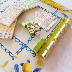 May include: A mahjong game set featuring white tiles with green and yellow lemon illustrations, a yellow tile rack, and instruction cards labeled Lemon Lime. The game board has a white surface with a blue scalloped border and yellow lemon motifs.