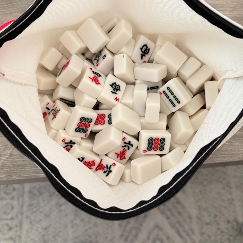 May include: A white canvas bag filled with numerous white Mahjong tiles. The tiles feature red and black symbols and numbers. The bag has a black zipper and a red accent.