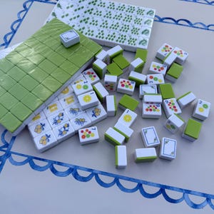 May include: A set of white and light green mahjong tiles featuring colorful fruit illustrations, including lemons, limes, and oranges. Some tiles are arranged in a rectangular block, while others are scattered, displaying numbers and fruit patterns.