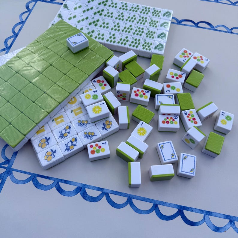 May include: A set of white and light green mahjong tiles featuring colorful fruit illustrations, including lemons, limes, and oranges. Some tiles are arranged in a rectangular block, while others are scattered, displaying numbers and fruit patterns.