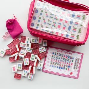 May include: A pink Mahjong set with white tiles featuring colorful symbols and numbers. The set includes a zippered case, a small drawstring bag, dice, and a pink card with the text "Glitter Sparkle".