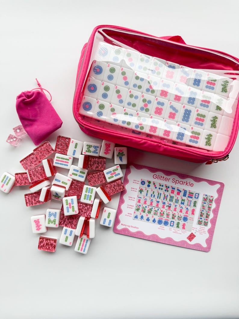 May include: A pink Mahjong set with white tiles featuring colorful symbols and numbers. The set includes a zippered case, a small drawstring bag, dice, and a pink card with the text "Glitter Sparkle".