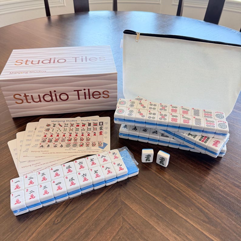 May include: A complete Mahjong set with white tiles featuring red and black symbols, stacked on a wooden table. The set includes a box labeled "Studio Tiles", a white zippered bag, and playing cards. The tiles have blue edges.