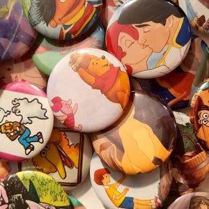 May include: A collection of circular pin-back buttons featuring various colorful cartoon characters, including Winnie the Pooh, Piglet, Ernie, Cookie Monster, Count von Count, and scenes from animated films, all arranged in a dense, overlapping pile.
