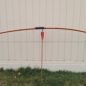 May include: A traditional wooden bow with a drawn bowstring and a red and white fletched arrow. The bow is light brown with black accents and is set against a white fence and green grass.