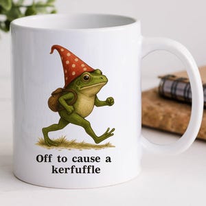 May include: White ceramic mug featuring a whimsical illustration of a green frog wearing a red and white polka-dot hat and carrying a backpack. The text "Off to cause a kerfuffle" is printed below the frog. A fun, novelty item.