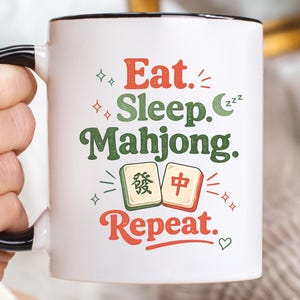 May include: White ceramic mug with a black handle and rim, featuring the text "Eat. Sleep. Mahjong. Repeat." in red and green. The mug also displays two Mahjong tiles. A fun gift for Mahjong players.