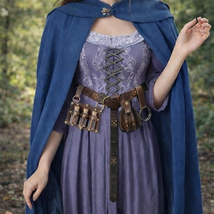 Peut inclure: Une cape bleue avec un fermoir décoratif est portée sur une robe lavande avec un corsage de style corset. Une ceinture en cuir marron avec des pochettes et des fioles est portée à la taille, créant une esthétique fantastique ou médiévale.