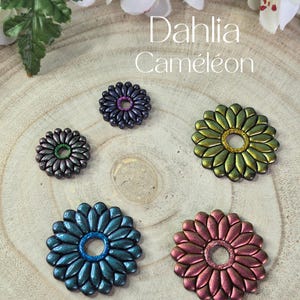 Peut inclure: Cinq pendentifs en forme de fleur Dahlia Caméléon de différentes couleurs, dont le bleu, le vert, le violet et le rose, sont présentés sur une surface en bois. Les pendentifs ont un aspect métallique et un centre scintillant. Le texte "Dahlia Caméléon" est visible.