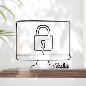 May include: Black metal sculpture of a computer monitor with a padlock icon on the screen. The name "Jackie" is written in cursive at the base of the monitor. The sculpture is displayed on a wooden surface.