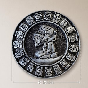 May include: A circular wall plaque featuring a silver-toned relief of a kneeling figure surrounded by a border of intricate glyphs. The plaque has a diameter of 20 cm as indicated by the measurement graphic. The design is set against a dark background.
