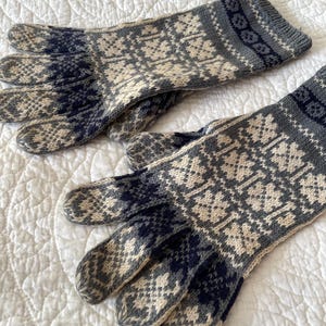 May include: A pair of knitted gloves with a geometric pattern in shades of gray, navy, and cream. The gloves are lying on a white, quilted surface. The gloves appear to be made of wool.