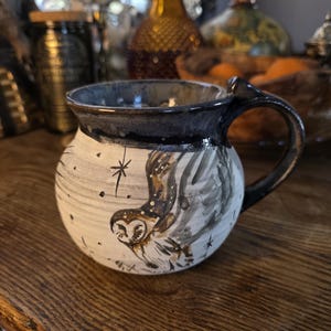 May include: A handmade ceramic mug with a dark blue rim and handle. The mug features a white base with a hand-painted design of an owl in flight, stars, and a textured background. The mug is approximately 4 inches tall and 4 inches in diameter.