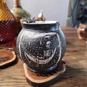 May include: Black ceramic ashtray with a white illustration of a skeleton relaxing in a hammock under string lights. The ashtray sits on a wooden coaster with a tree design. Decorative bottles are in the background.
