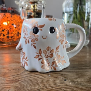 Peut inclure: Une tasse en céramique blanche en forme de fantôme avec une anse, décorée de motifs floraux couleur cuivre. La tasse a des yeux noirs et une bouche souriante. La tasse est posée sur une surface en bois.
