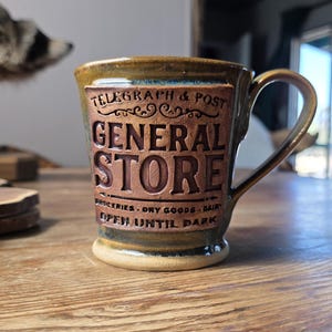 May include: A ceramic mug with a brown and green glaze. The mug features the words "TELEGRAPH & POST GENERAL STORE" in a raised, vintage-style design. Additional text reads "GROCERIES - DRY GOODS - DAIRY OPEN UNTIL DARK."