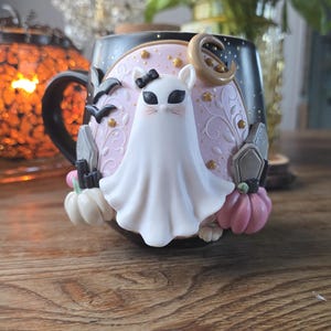 May include: A black ceramic Halloween mug featuring a white ghost cat design. The mug has a black handle and is decorated with bats, pumpkins, and a crescent moon. The background is a light pink color with gold accents.