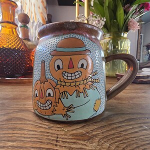 May include: A ceramic mug with a whimsical design featuring a scarecrow and a pumpkin. The mug has a brown handle and rim, with a light blue background dotted with white circles. The scarecrow wears a hat and has a stitched mouth.