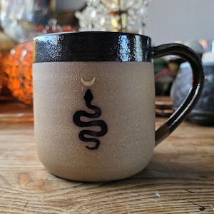 May include: A beige ceramic mug with a black rim and handle. The mug features a black snake design with a crescent moon above it. The mug is on a wooden surface.