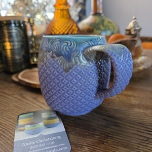 May include: A handmade ceramic mug with a textured, diamond pattern in shades of blue and purple. The handle is also textured. The mug has a unique glaze with a gradient of colors. A business card is visible with contact information.