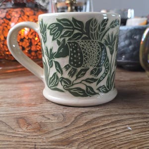 May include: Cream-colored ceramic mug with a green armadillo design. The armadillo is surrounded by green foliage. The mug has a curved handle and a slightly flared base. The mug is sitting on a wooden surface.