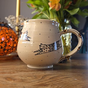 May include: A beige ceramic mug with a rounded shape and a handle. The mug features black illustrations of flying creatures and small black dots. The handle has a decorative pattern. The mug is on a wooden surface.