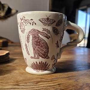 May include: Cream-colored ceramic mug with a brown hare illustration, wheat, and floral accents. The mug has a curved handle and a flared base. The design is repeated around the mug. A moth and feather details are also included in the design.