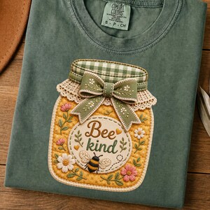 Peut inclure: T-shirt vert sauge avec un motif de bocal décoratif. Le bocal a un fond jaune, un couvercle à carreaux verts et blancs et un nœud vert. Le motif comprend les mots "Bee kind" et une illustration d'abeille.
