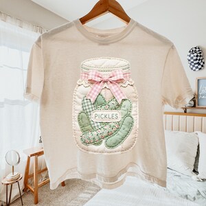 A beige t-shirt with a detailed embroidered design of a jar filled with pickles, featuring a pink bow and the word "PICKLES" printed on it.