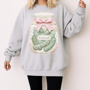 A person wearing a gray sweatshirt with a large embroidered image of a jar filled with pickles.