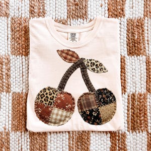 May include: Pale pink t-shirt with a large cherry design. The cherries are made of patchwork fabric with various patterns, including leopard print, plaid, and floral designs. The stems and leaves are brown.