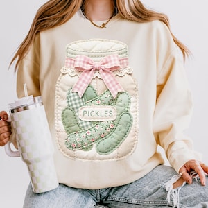 A person wearing a sweater with a large embroidered jar of pickles on it, holding a drink in a cup.