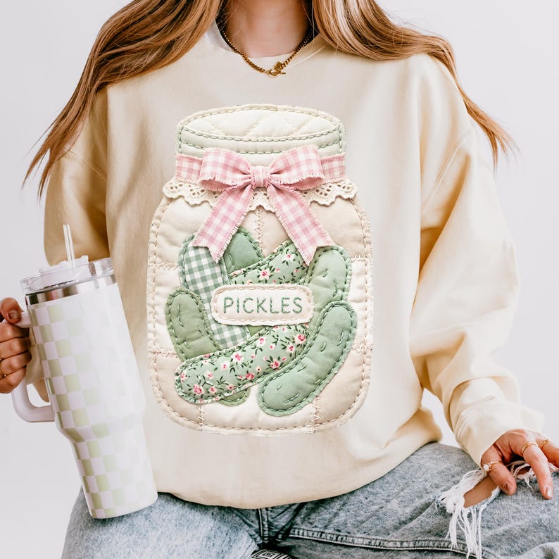 A person wearing a sweater with a large embroidered jar of pickles on it, holding a drink in a cup.