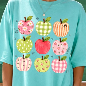 Könnte beinhalten: Ein hellblaues T-Shirt mit einem Grafikdruck von neun Äpfeln in einem Raster. Die Äpfel zeigen verschiedene Muster, darunter weiße Gänseblümchen, grünes Vichy-Karo, orangefarbene Streifen, weiße Punkte, einfarbiges Rot, rosa Blumen, rosa Streifen mit Herzen und rosa Vichy-Karo.