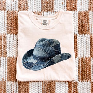 May include: A light peach-colored t-shirt featuring a denim patchwork cowboy hat. The hat is made of various shades of blue denim and has a stitched brim. The shirt has a Comfort Colors tag.