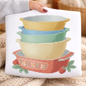 May include: A white cushion with a cross-stitch design of stacked mixing bowls in pastel shades: yellow, blue, green, and peach. The bottom bowl has a strawberry and floral pattern. The cushion is held by a person.