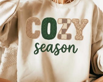 May include: A beige sweatshirt with the words "COZY season" in a mix of knitted and script fonts. The word "COZY" is in a combination of brown, green, and checkered patterns. The word "season" is in a green script font.
