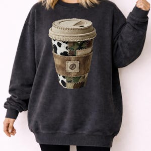 May include: Dark grey sweatshirt with a coffee cup design. The cup features patches of cow print, camouflage, and brown fabric. A brown leather-like band with a coffee bean emblem wraps around the cup. The lid is beige with stitched detailing.