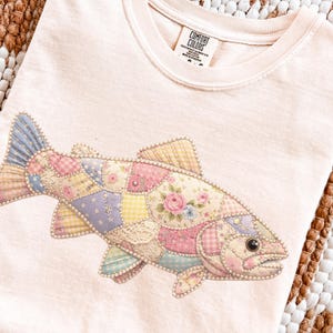 May include: A light pink t-shirt featuring a colorful patchwork fish design. The fish is made of various patterned fabrics, including floral, polka dot, and gingham, with pearl-like embellishments. The shirt has a crew neck and short sleeves.
