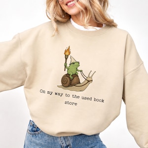 Può includere: Felpa beige con un'illustrazione di una rana verde con un cappello da mago che cavalca una lumaca e tiene una torcia. Il testo "On my way to the used book store" è stampato sotto la grafica.